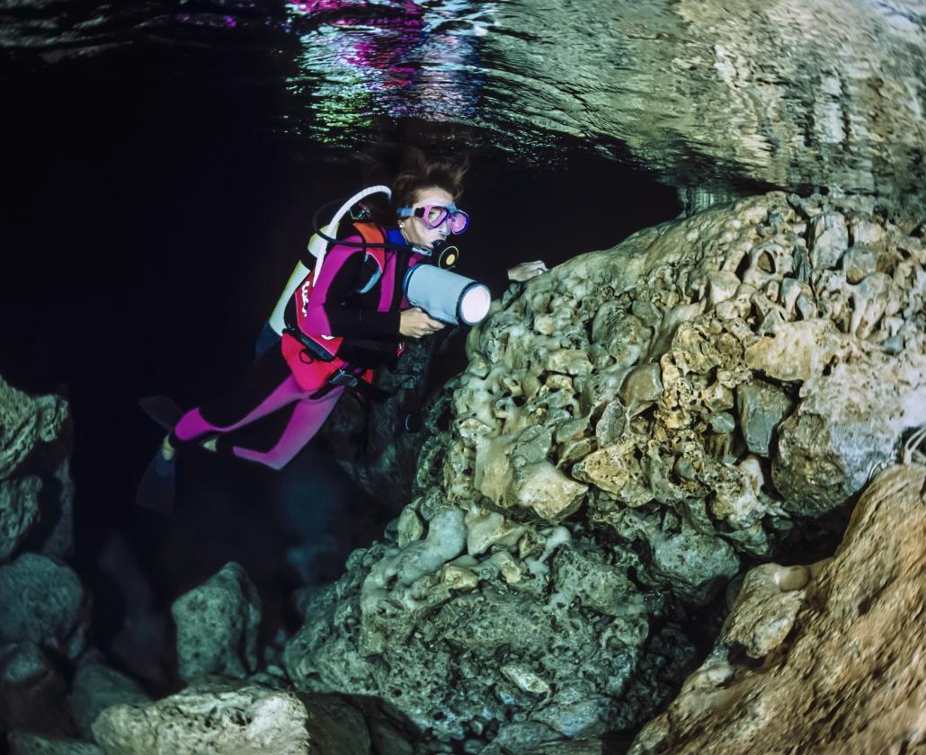 Subacquea esplora una grotta marina con torcia, indossando muta rosa e attrezzatura subacquea.