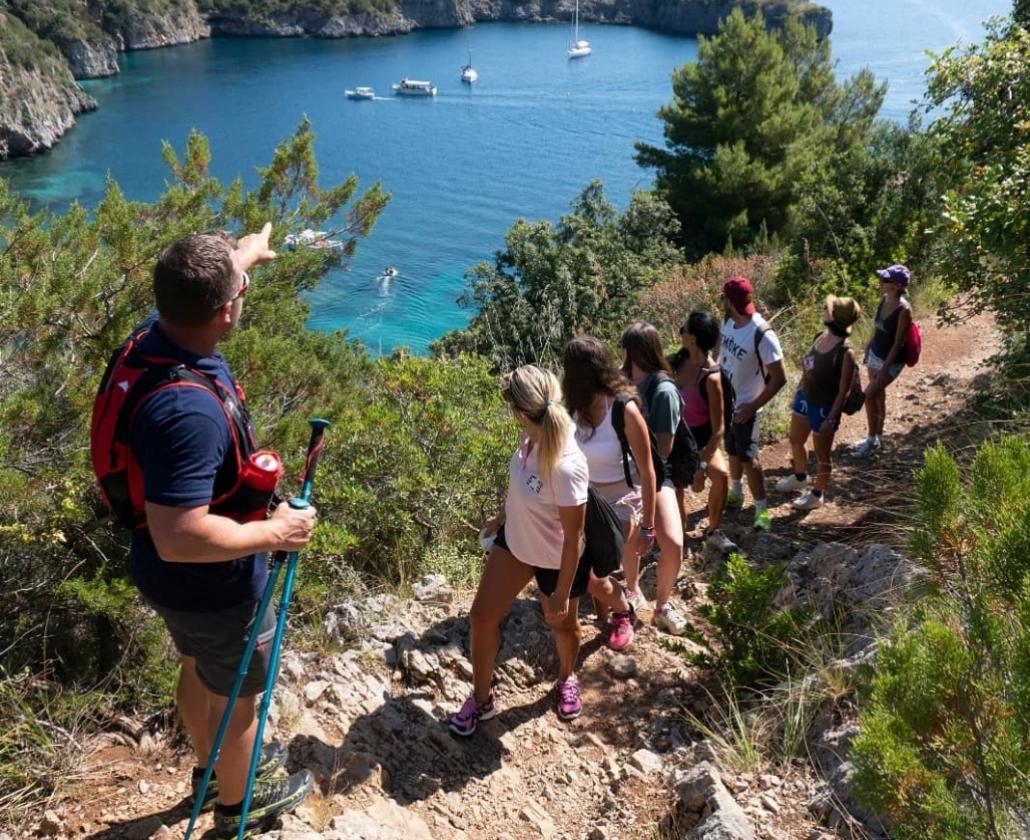 Gruppo di escursionisti ammirano il panorama costiero con barche e mare blu.