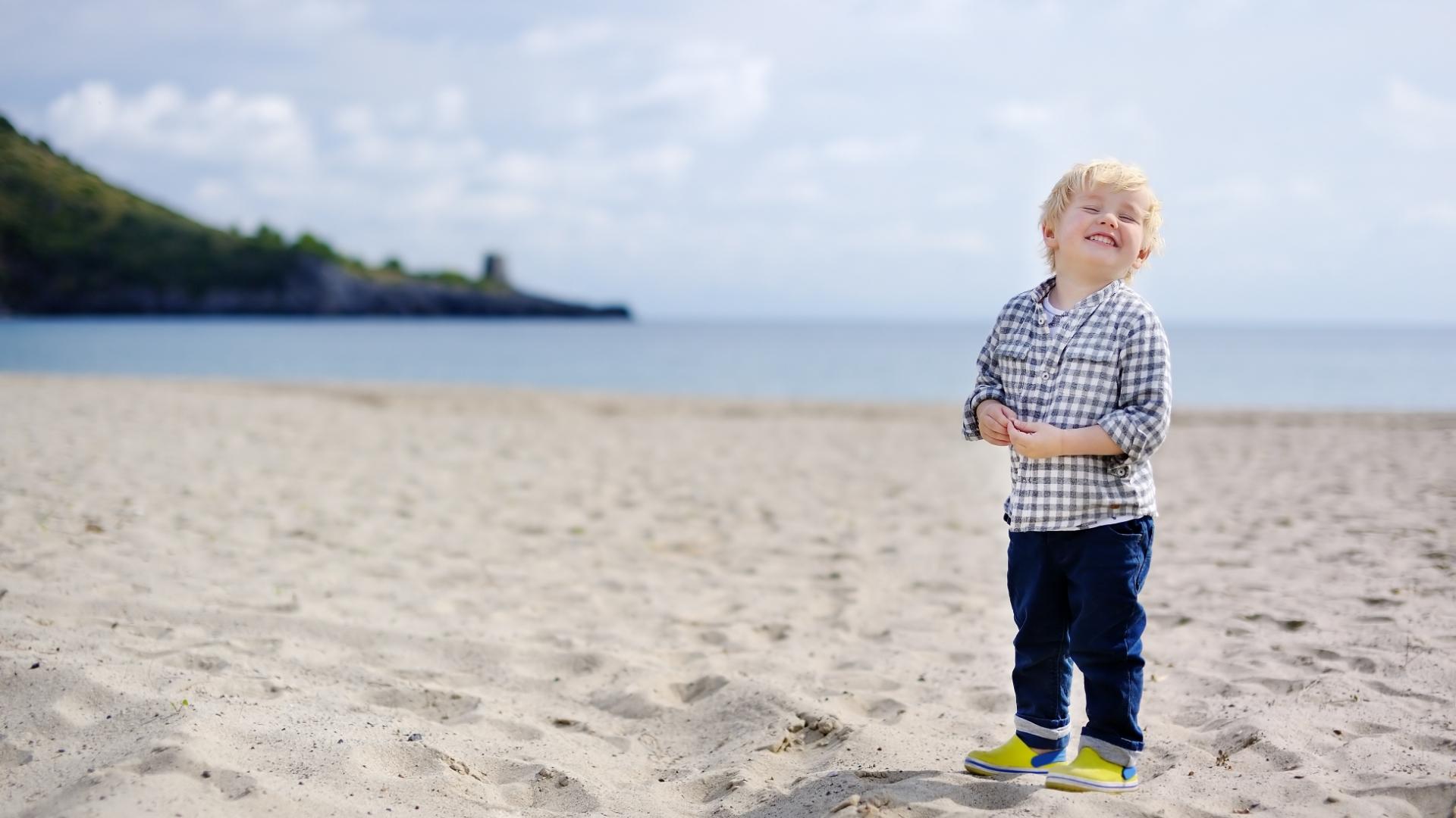 Bambino felice sulla spiaggia con camicia a quadri e scarpe gialle.