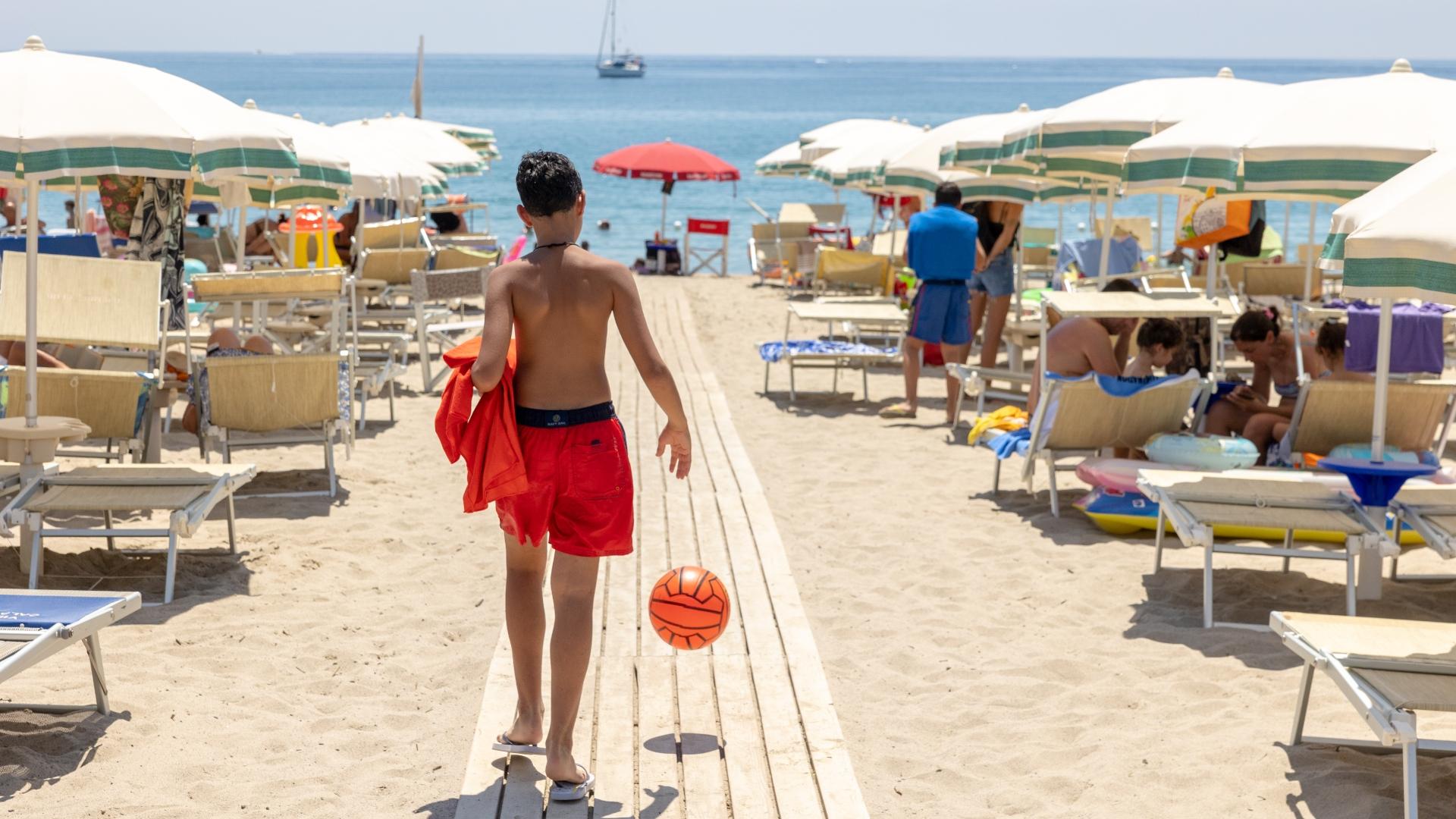 Ragazzo in spiaggia con pallone, cammina verso il mare tra ombrelloni e sdraio.