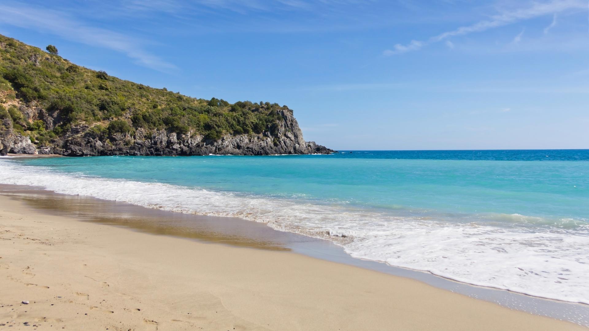 Spiaggia sabbiosa con mare turchese e scogliera verde sotto un cielo sereno.