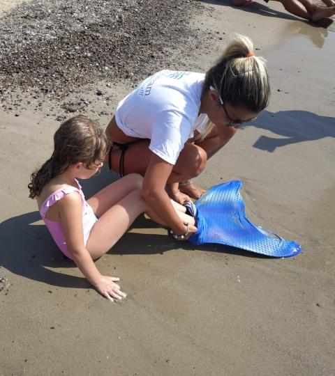 Bambina in spiaggia indossa una coda da sirena con l'aiuto di un adulto.