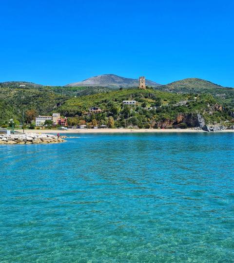 Spiaggia con acqua cristallina, colline verdi e una torre storica sullo sfondo.