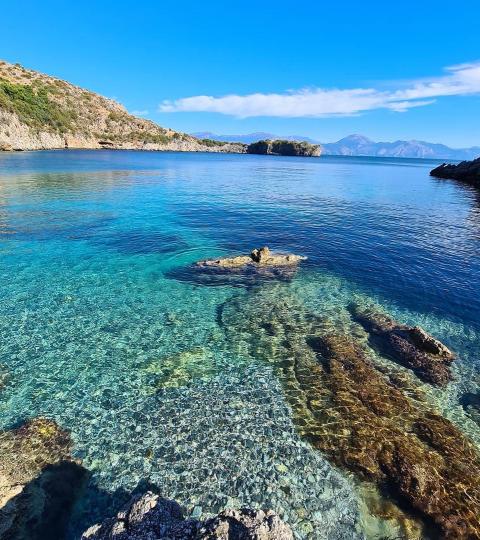 Mare cristallino con rocce, cielo azzurro e colline sullo sfondo.