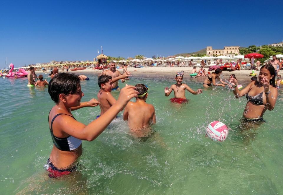 Persone giocano a pallone in acqua, sotto un cielo limpido, in una spiaggia affollata.